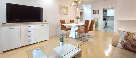 Una sala de estar moderna con un televisor de pantalla plana, una mesa de centro de vidrio y un sofá. Se observa un comedor con una mesa blanca y sillas marrones. La cocina es visible al fondo.