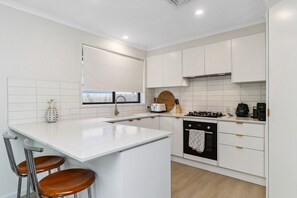 A modern kitchen with white cabinetry, a central island with stools, a stove, and a window with a view. - Bay Breeze House  - Capel Sound (Capel Sound)