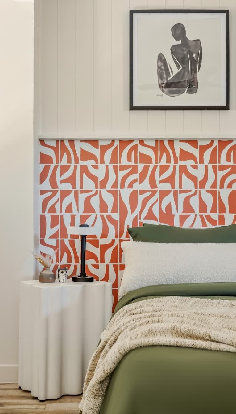 Une chambre avec un papier peint à motifs, une œuvre d’art encadrée au mur, une table de chevet avec une lampe et un lit avec une literie verte.