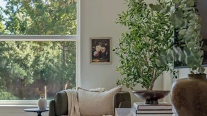 Una habitación acogedora con un ventanal grande, una silla verde, un aparador blanco y una planta en maceta.