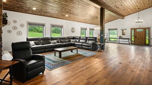 A spacious living room with a large black leather sectional sofa, a wooden coffee table, and a colorful rug. The room features a high wooden ceiling, large windows with a view of greenery, and a door leading outside. - The Zoo Barn! Pool, Hot Tub, Bball/Pkbl, Theater (Spring Green)