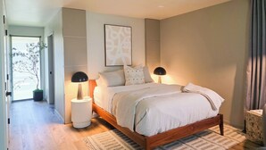 A bedroom with a wooden bed, two bedside lamps, a large abstract painting, and a view of a tree through an open door. - Walk to River: Myrtle Creek Studio Near I-5 (Myrtle Creek)