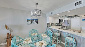 Ein Essbereich mit Glastisch und türkisfarbenen Stühlen, eine Küche mit weiß gefliestem Fliesenspiegel und einem Deckenventilator.