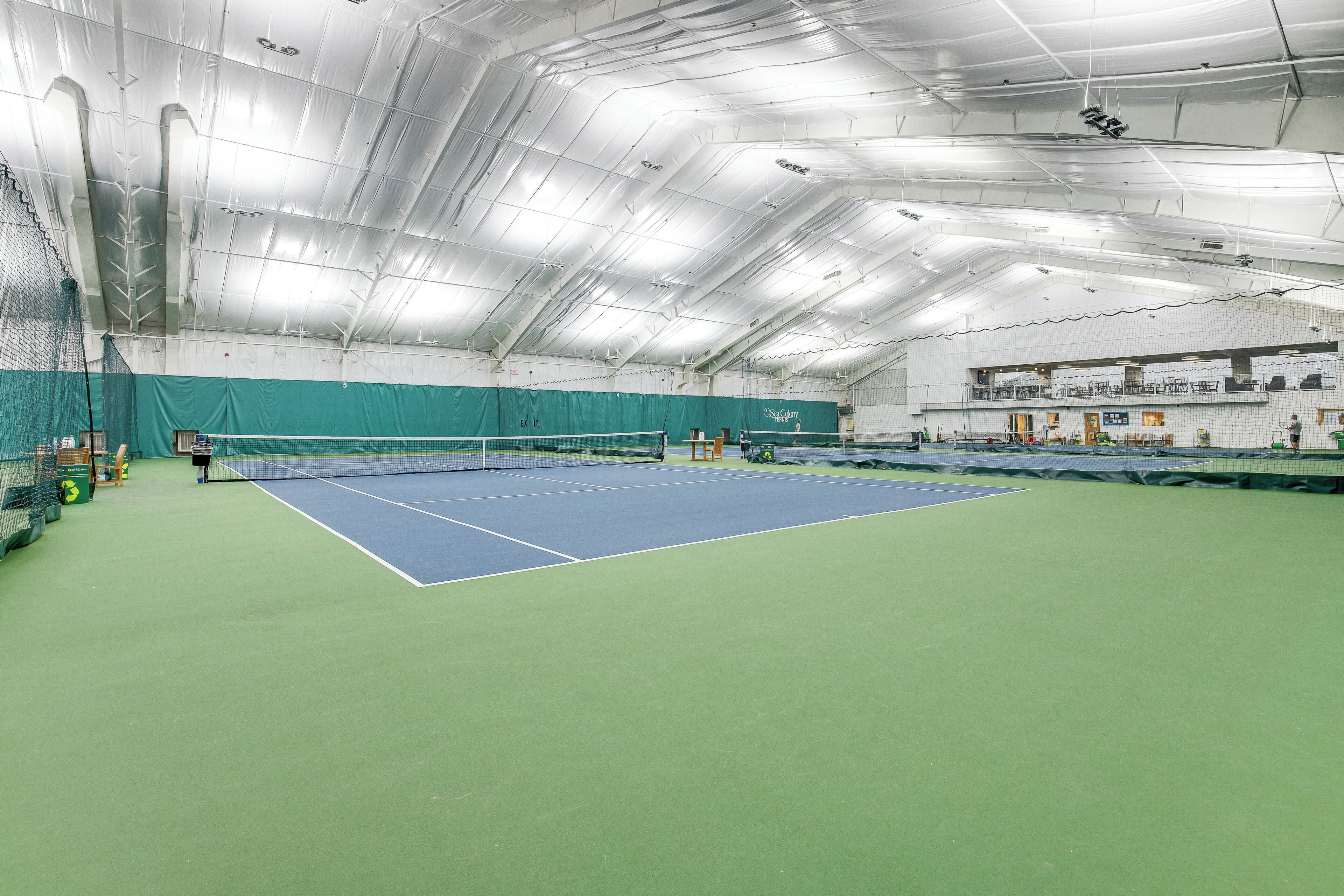 En indendørs tennisbane med net, grønne og blå spilleflader og et højt loft med lysarmaturer.