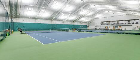 En indendørs tennisbane med net, grønne og blå spilleflader og et højt loft med lysarmaturer.