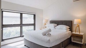 A modern bedroom with a large window, a grey bed, two bedside tables with lamps, and a wooden nightstand. - The Carlton Residences (Melbourne)