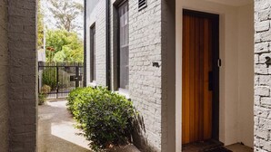 A narrow pathway leading to a door with a wooden panel, flanked by a brick wall and a small bush. - The Carlton Residences (Melbourne)