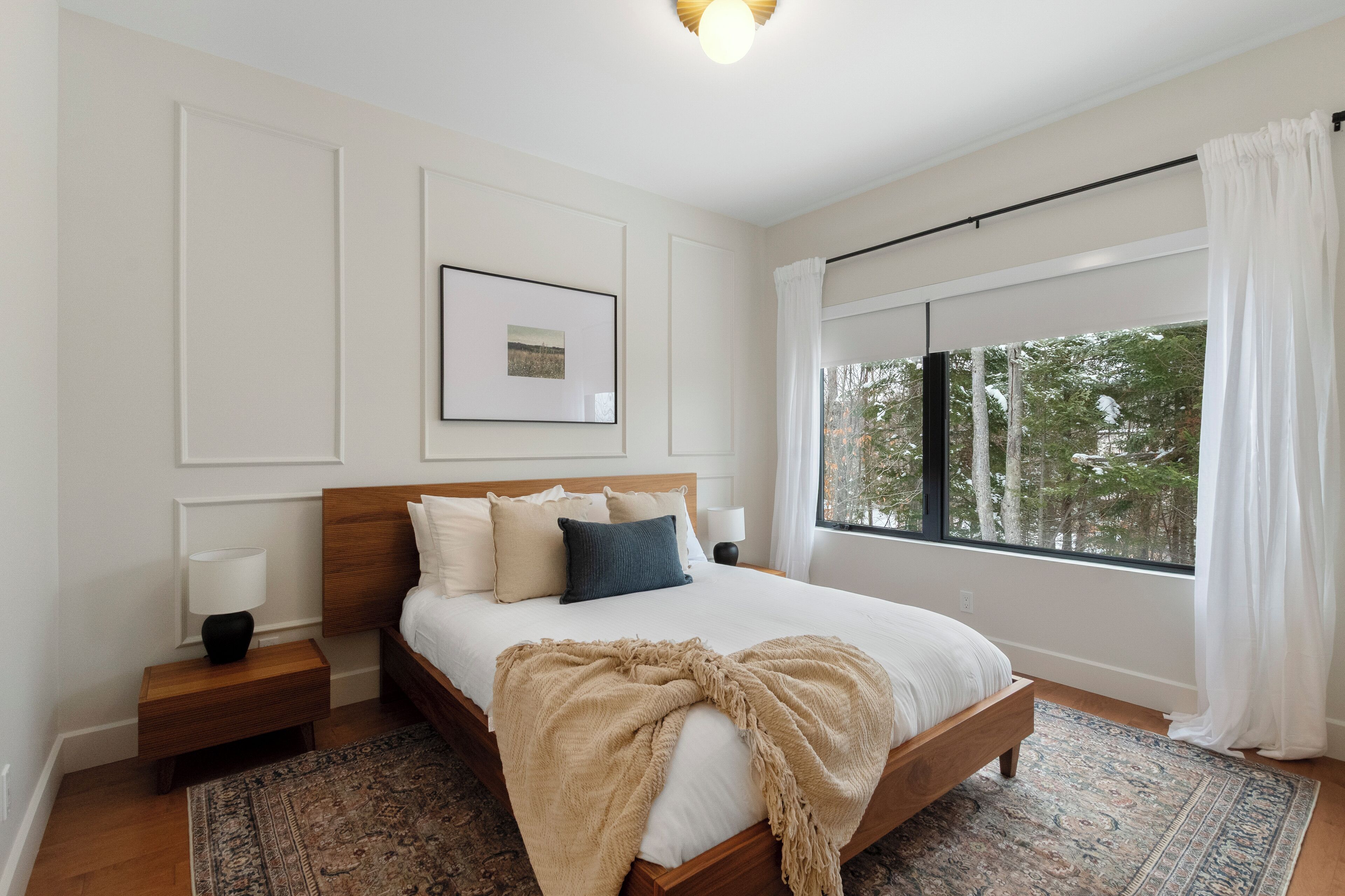 A bedroom with a wooden bed, white walls, a framed picture, a nightstand with a lamp, and a large window with a view of trees.