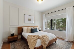 A bedroom with a wooden bed, white walls, a framed picture, a nightstand with a lamp, and a large window with a view of trees. - L'Orignal et L'Érable by Tremblant Platinum (La Conception)