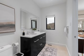 A modern bathroom with a black vanity, white fixtures, a large mirror, and a view of trees through a window. - L'Orignal et L'Érable by Tremblant Platinum (La Conception)