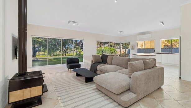 A spacious living room with a large sectional sofa, a black coffee table, and a fireplace. There are large windows offering views of a garden and a kitchen area with a countertop and sink.