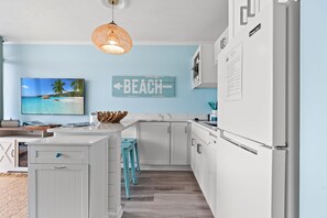 A kitchen with white cabinetry, a teal island, a hanging light fixture, a wall-mounted television, and a 'BEACH' sign. - Oceanfront Condo*Walkout Patio Beach Access*Steps to the Sea* (North Myrtle Beach)