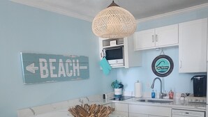 A kitchen with a white island, teal bar stools, a woven pendant light, and a 'Beach' sign on the wall. - Oceanfront Condo*Walkout Patio Beach Access*Steps to the Sea* (North Myrtle Beach)