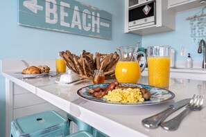 Preparación de desayuno con cruasanes, muffins, tocino, huevos, salchichas y jugo sobre una encimera blanca.