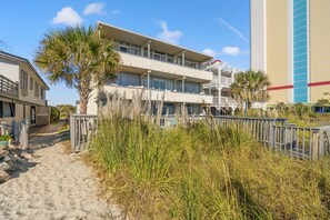 A beachfront hotel with a sandy walkway, palm trees, and a building with multiple balconies. - Oceanfront Condo*Walkout Patio Beach Access*Steps to the Sea* (North Myrtle Beach)