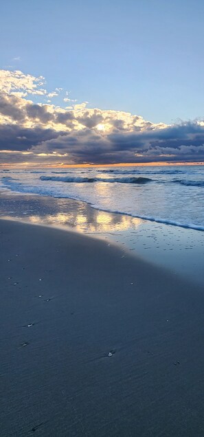 A beach with wet sand and gentle waves, under a partly cloudy sky with the sun setting or rising. - Oceanfront Condo*Walkout Patio Beach Access*Steps to the Sea* (North Myrtle Beach)