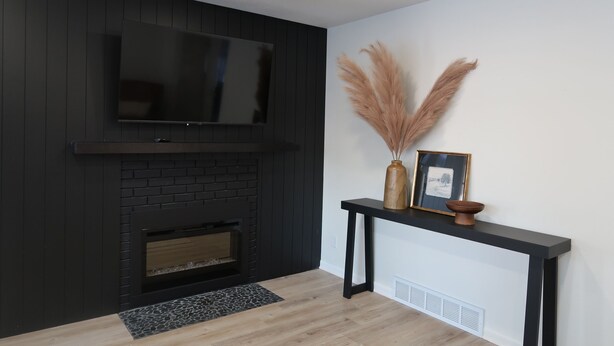 Una sala de estar moderna con chimenea de ladrillo negro, un televisor de pantalla plana montado en la pared y una mesa consola negra con adornos.