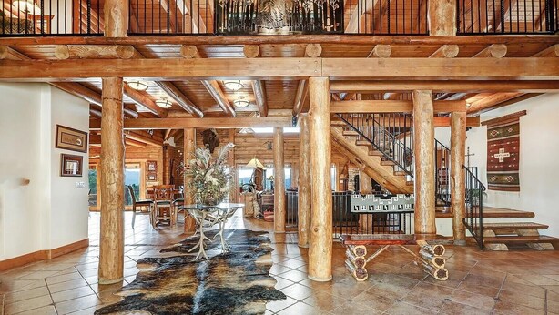 A spacious interior with a high ceiling, wooden beams, a staircase, and a dining area with a table and chairs. - Breathtaking Log Home w/Panoramic Mountain Views, Hot Tub, 2 Game rooms, 6 bdrms (Angel Fire)