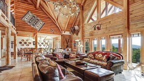 A spacious living room with high ceilings, wooden beams, and large windows offering a view of the outdoors. - Breathtaking Log Home w/Panoramic Mountain Views, Hot Tub, 2 Game rooms, 6 bdrms (Angel Fire)