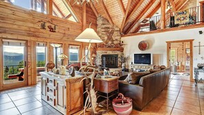 A spacious living room with a fireplace, wooden ceiling, and a large window overlooking a scenic view. - Breathtaking Log Home w/Panoramic Mountain Views, Hot Tub, 2 Game rooms, 6 bdrms (Angel Fire)