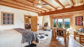 A rustic cabin interior with a large bed, a fireplace, a dining area, and a view of the mountains. - Breathtaking Log Home w/Panoramic Mountain Views, Hot Tub, 2 Game rooms, 6 bdrms (Angel Fire)