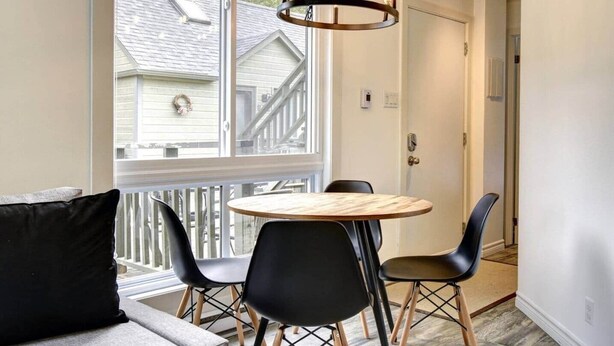 A dining area with a round wooden table and four black chairs, a chandelier, and a view of a house through large windows.