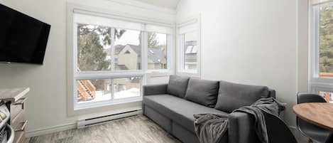 Een moderne woonkamer met een grijze bank, een flatscreen televisie en een eethoek met een houten tafel en stoelen.