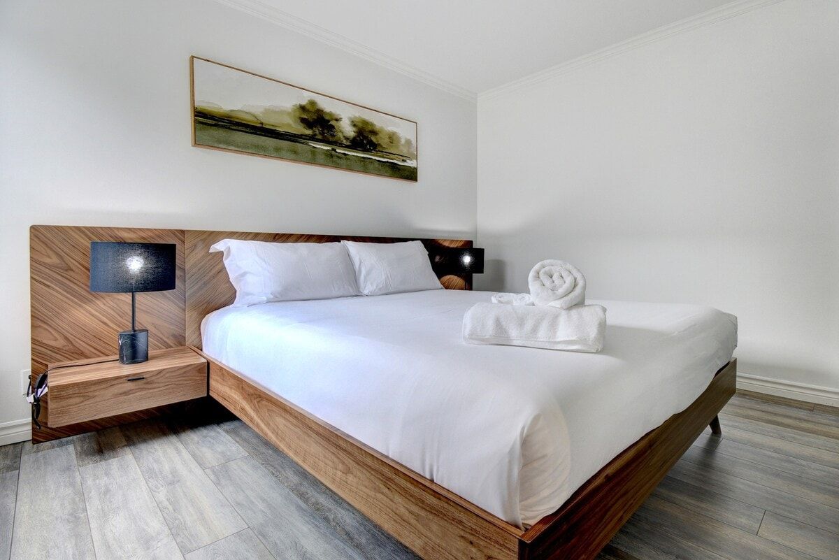 Moderni hotellihuone, jossa on puinen sängynrunko, valkoiset vuodevaatteet, yöpöytä lampun kanssa ja taulu seinällä.
