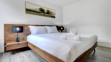 Moderni hotellihuone, jossa on puinen sängynrunko, valkoiset vuodevaatteet, yöpöytä lampun kanssa ja taulu seinällä.