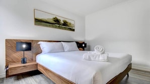 A modern hotel room with a wooden headboard, a single bed with white linens, a nightstand with a lamp, and a painting on the wall.