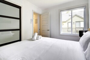 A hotel room with a bed, a window with a view, a door, and a wall-mounted light fixture.
