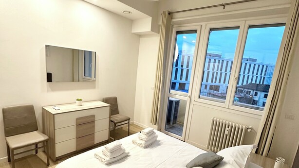 Ein modernes Schlafzimmer mit einem großen Bett, einem Schreibtisch, einem Stuhl und einem Fenster mit Blick auf die Stadt.