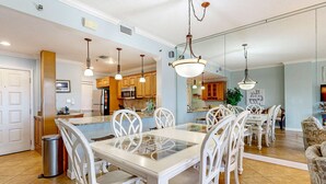 Una cocina amplia con electrodomésticos modernos, un comedor con mesa de vidrio y sillas blancas, y una sala de estar con sofá y televisor.