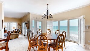 A dining area with a round table and chairs, a chandelier, and large windows with a view of the beach.