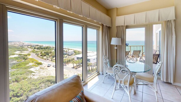 Un balcon avec une table et des chaises en verre, une vue sur la plage et une grande fenêtre avec des rideaux.
