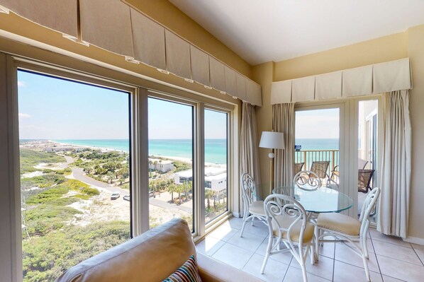 A balcony with a glass table and chairs, a view of the beach, and a large window with curtains.