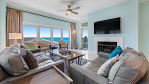 A spacious living room with a large window overlooking the ocean, a ceiling fan, a flat-screen TV, and a fireplace. - Exclusive 3BR Beachfront with Gulf Views (Miramar Beach)