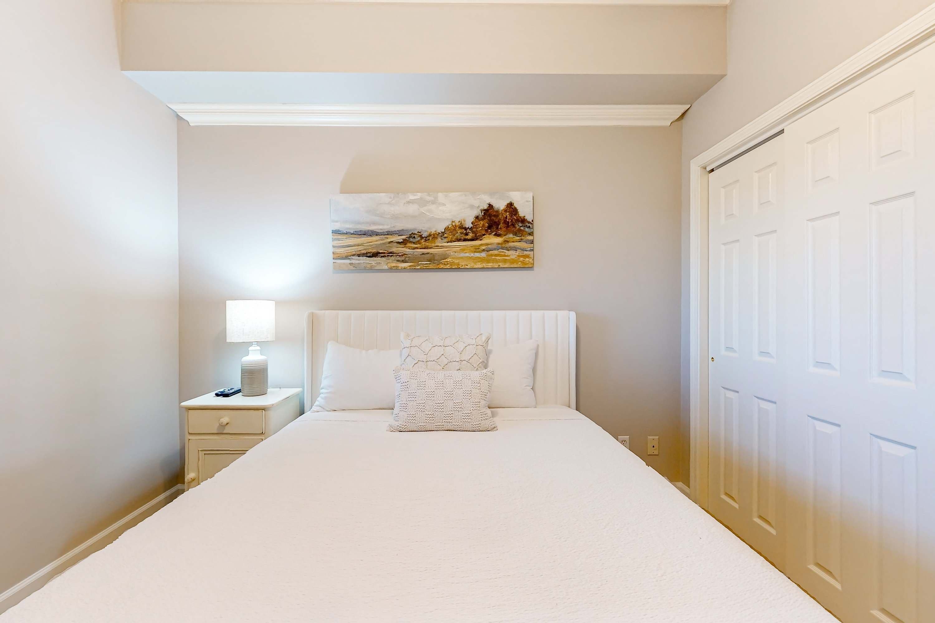 A bedroom with a bed, a nightstand, a lamp, a painting, and a door.