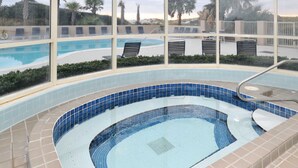 En swimmingpool med et bølgende design, omgivet af beige fliser og et glasvindue med udsigt over poolområdet og palmer.