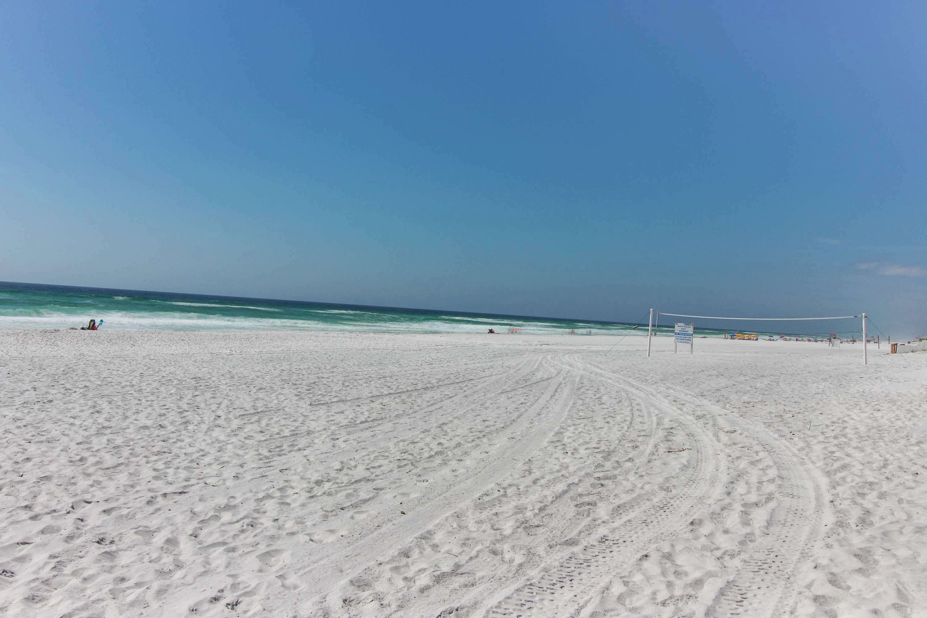 A sandy beach with footprints leading to the water, a volleyball net, and a clear blue sky.