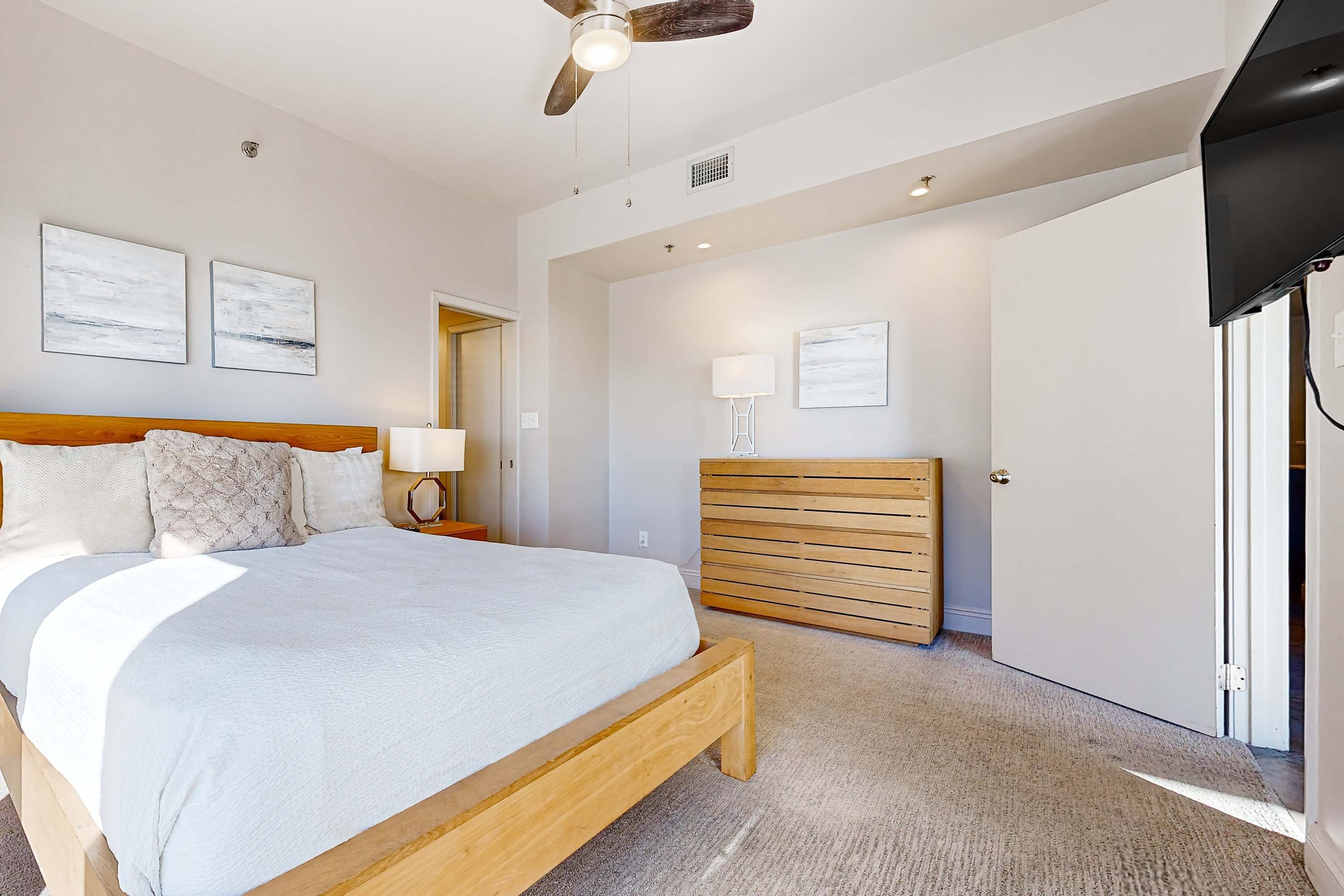 A bedroom with a bed, a ceiling fan, a bedside table with a lamp, a wooden headboard, a wall-mounted TV, and a door.