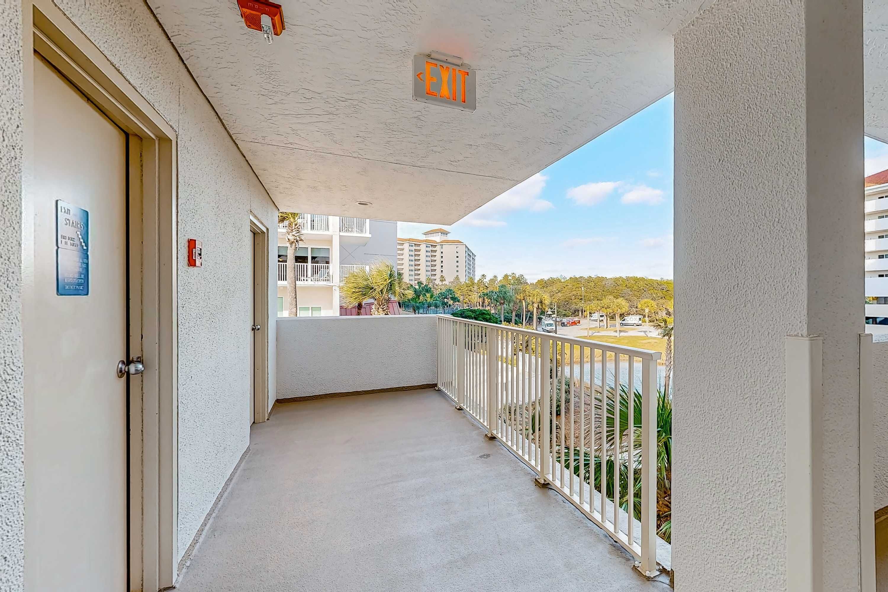 A balcony with a white railing, a door with a sign indicating the exit, and a view of buildings and trees in the background.