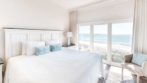 Quarto de hotel com uma cama grande, mesa de cabeceira, uma cadeira e vista para a praia.