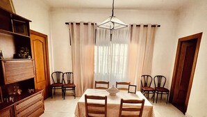 Comedor con mesa dispuesta para cuatro personas, rodeada de sillas, una lámpara de araña y cortinas.