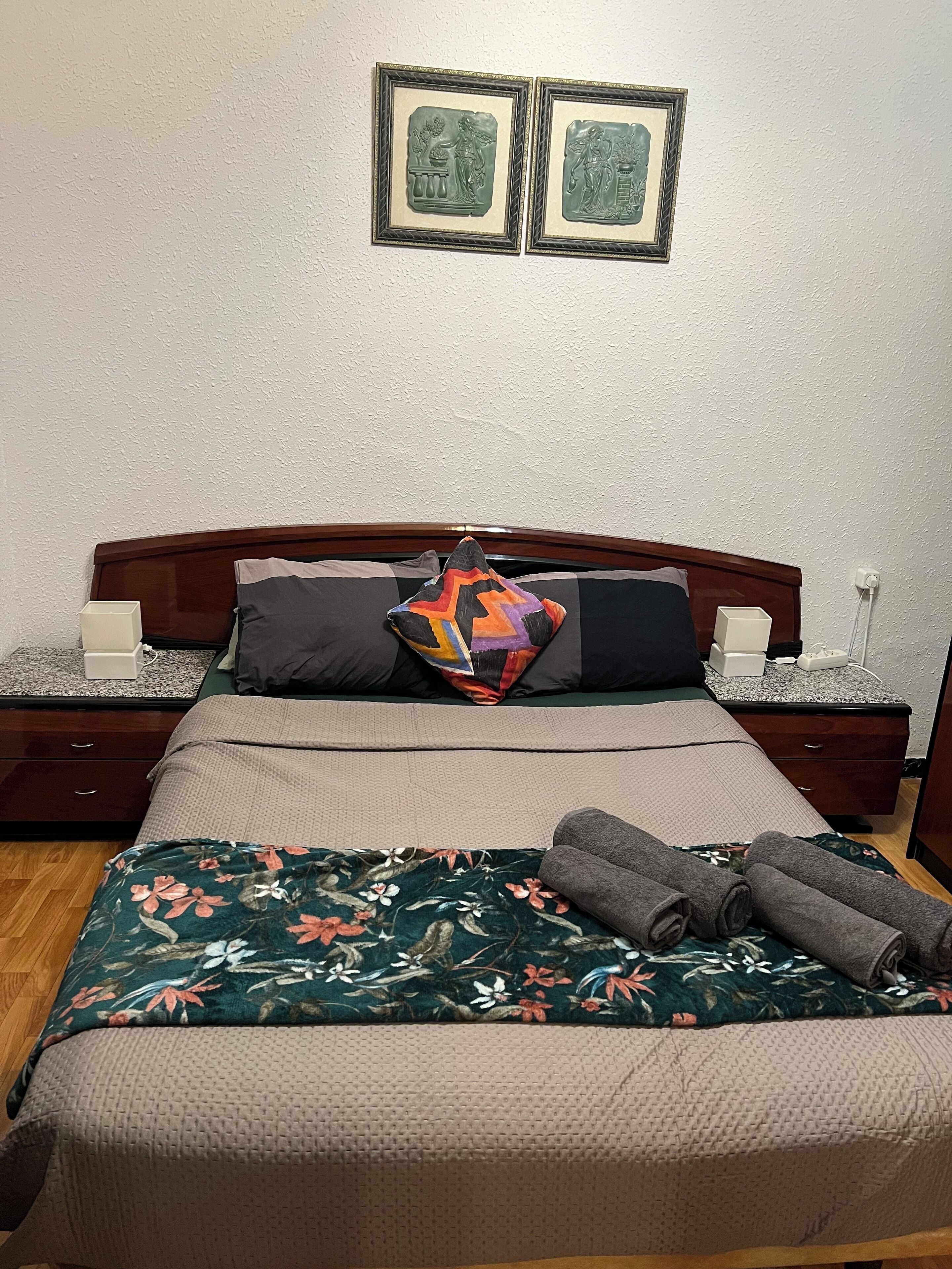Ein ordentlich bezogenes Bett mit floralem Überwurf, einem farbenfrohen geometrischen Kissen und aufgerollten Handtüchern seitlich. Zwei Nachttische mit Lampen und ein an der Wand hängendes Bild befinden sich über dem Bett.