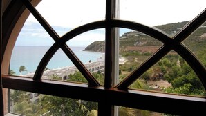 A view through a window with a geometric frame, overlooking a coastal area with a beach, a white building with arches, and a hilly landscape. - 5BR Villa, Pool & Little Bay Views (Little bay)