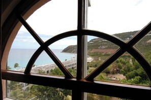 A view through a window with a geometric frame, overlooking a coastal area with a beach, a white building with arches, and a hilly landscape. - 5BR Villa, Pool & Little Bay Views (Little bay)