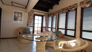 A spacious living room with a glass-top coffee table, wicker furniture, and large windows with blinds. - 5BR Villa, Pool & Little Bay Views (Little bay)