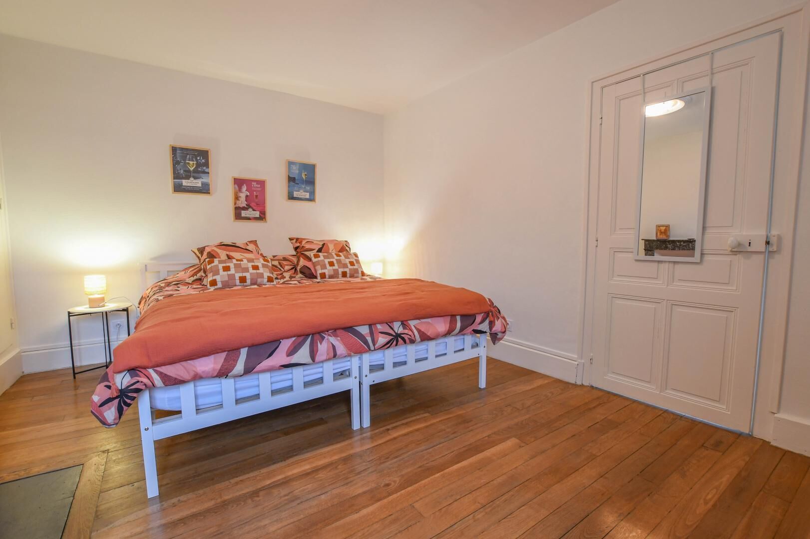 Een slaapkamer met een bed, houten vloeren, een nachtkastje met een lamp en een deur met een spiegel.