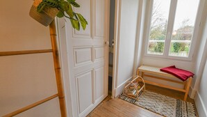 Een kamer met een houten vloer, een witte deur, een raam met uitzicht op groen, een klein bankje met een rode handdoek en een plant die aan het plafond hangt.